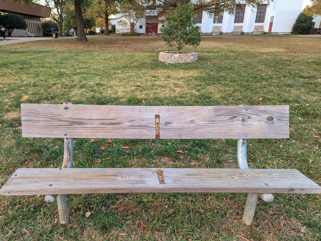 A faded wood bench with metal supports, set in a grassy yard outside the Greenbelt Library. A darker, un-faded patch of wood in the middle of the bench shows where an armrest was removed. Photo by Anna Bedford-Dillow.