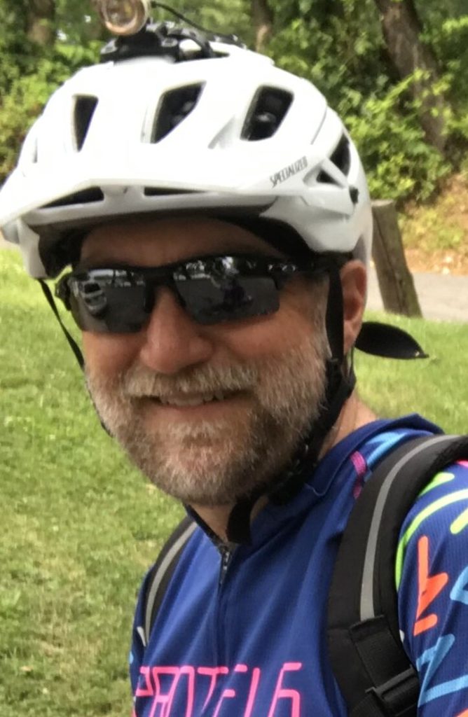 A light-skinned man wears a white bike helmet, sunglasses, and a blue shirt with bright designs. He has a close-trimmed pale beard and mustache. He is outside, with grass and a tree in the background.